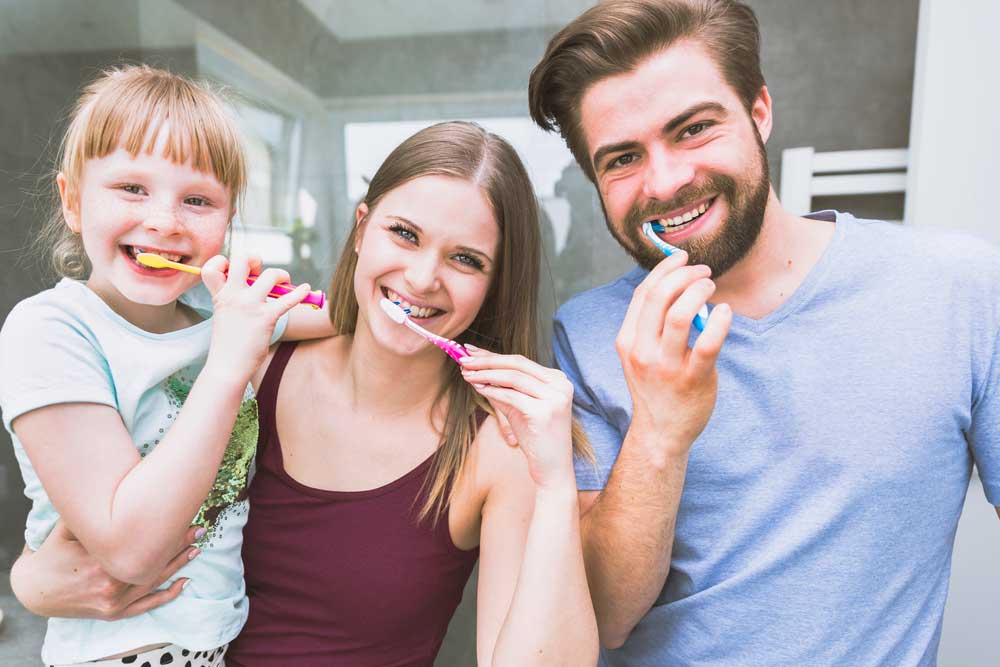 A family of three smiling and brushing their teeth together.