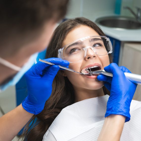 dentist in mask filling the patient's root canal while she is lying on dental chair in safety glasses under the medical lamp in clinic.